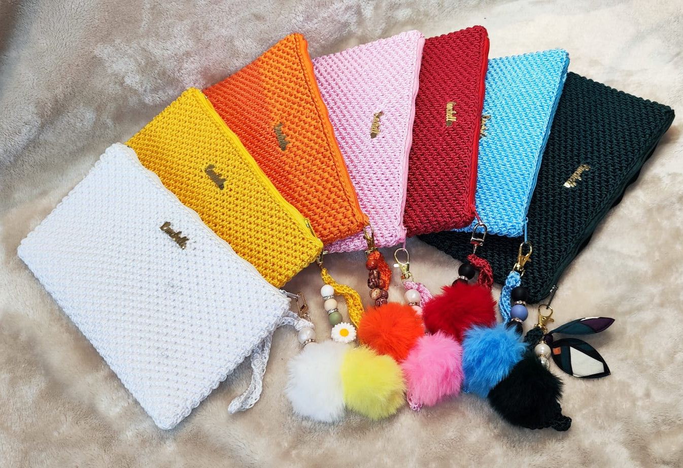 Full set of Crochella Luxe Clutches with gold logo and unique pom-pom keychains, styled on a cozy background.