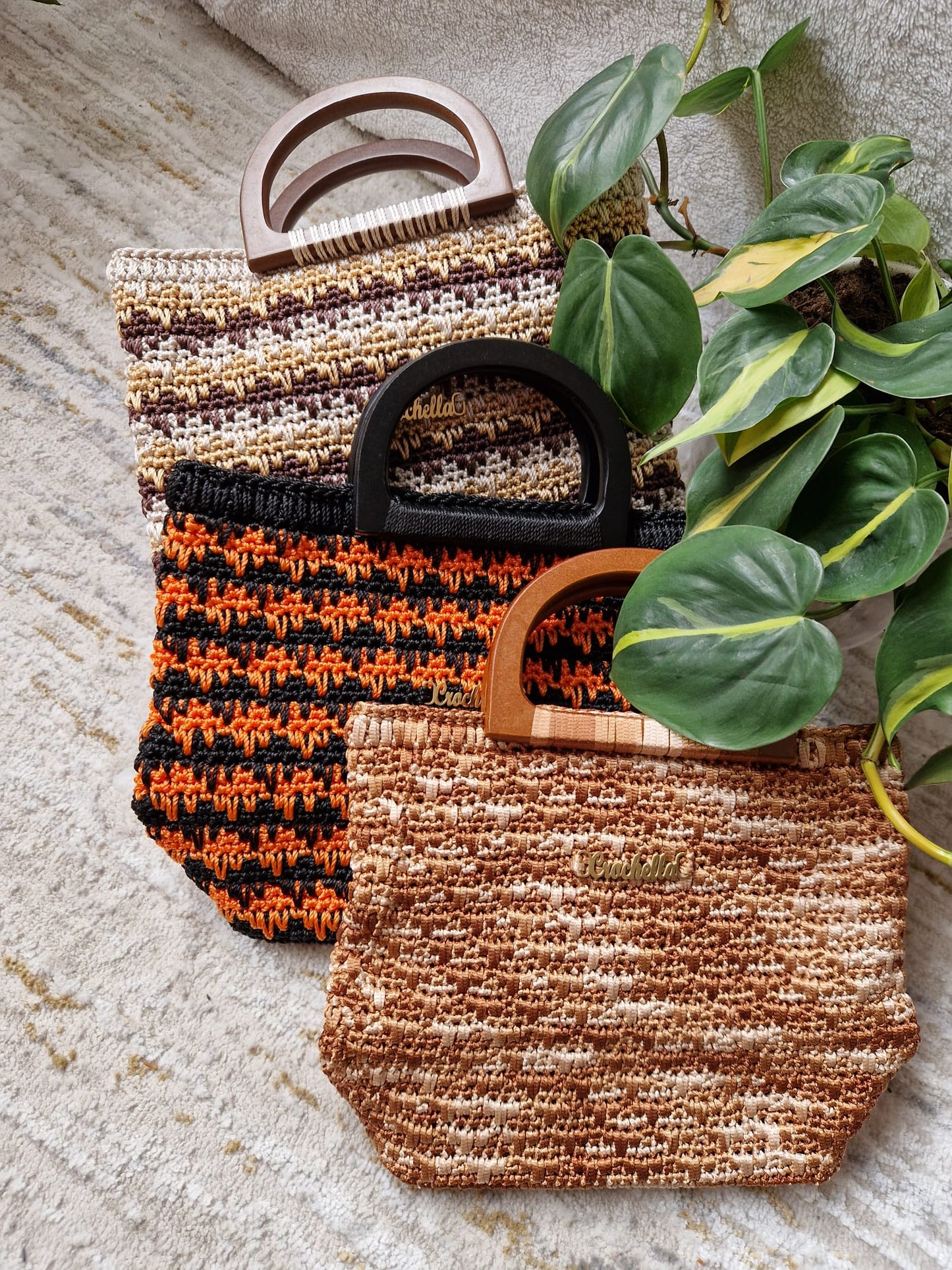 Three Ember Stripes Crochet Handbags in earthy tones, arranged with a leafy plant. Each features a different striped pattern and a wooden handle.