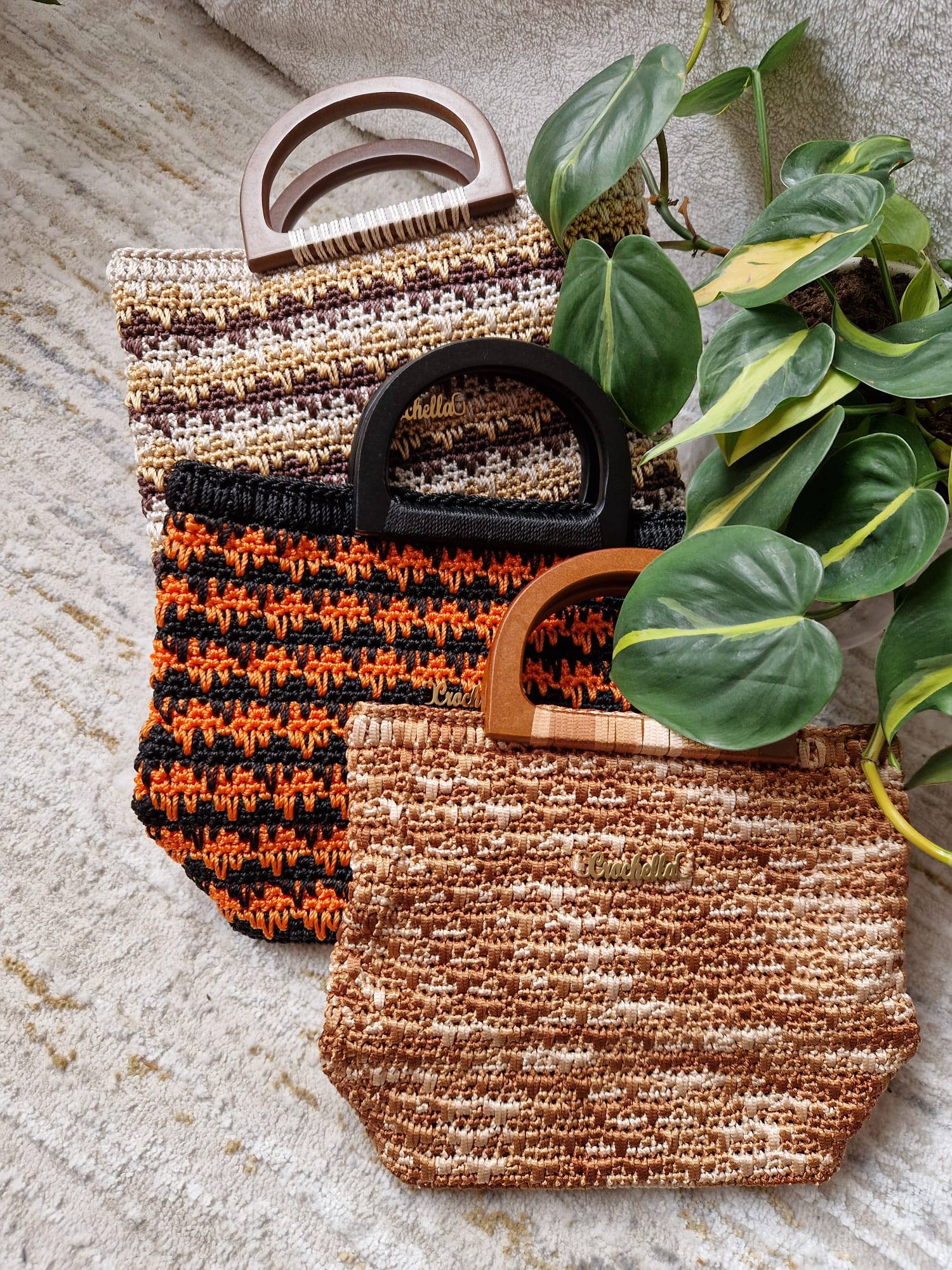 Three Ember Stripes Crochet Handbags in earthy tones, arranged with a leafy plant. Each features a different striped pattern and a wooden handle.