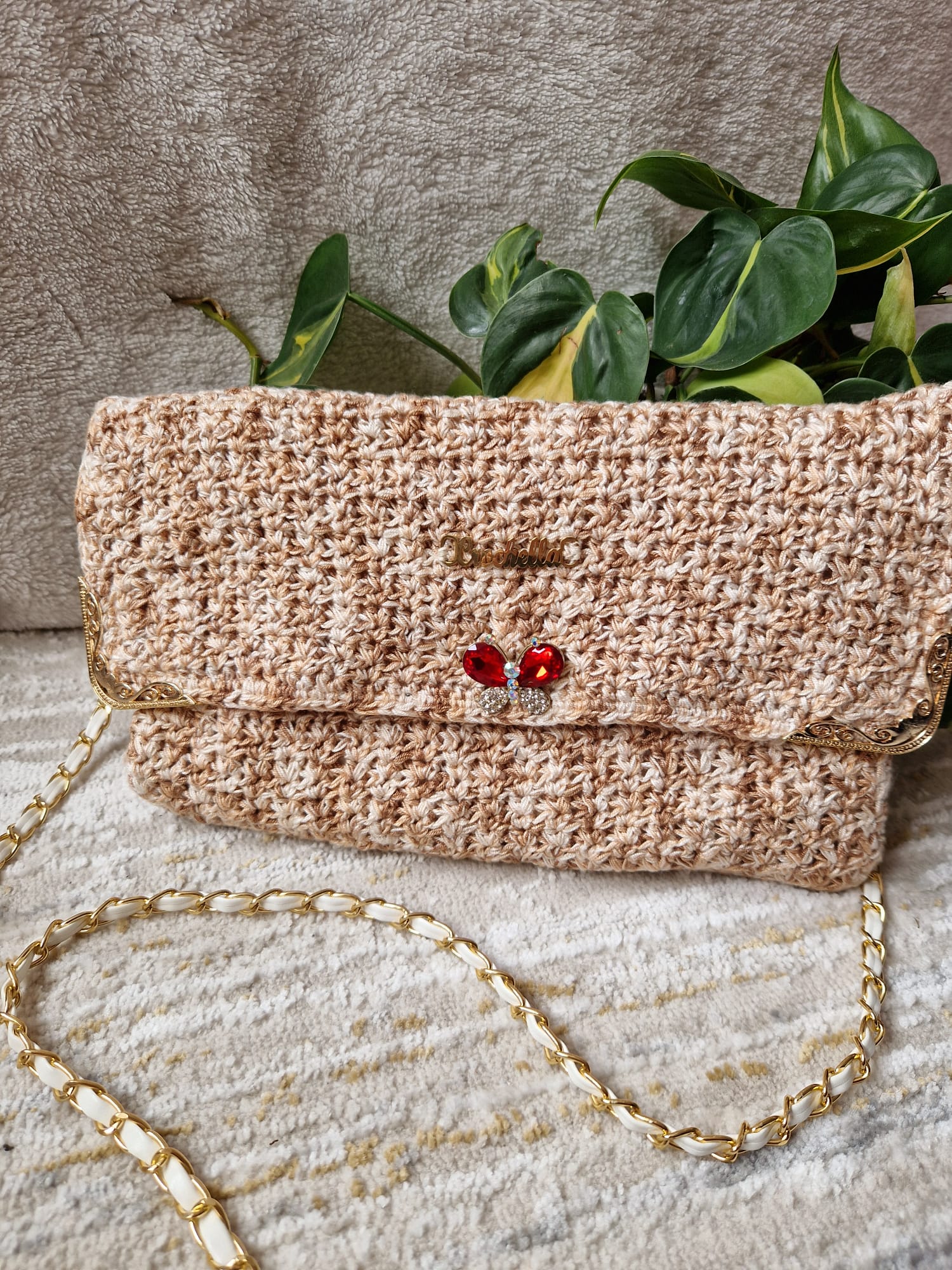 Golden Elegance Crochet Clutch with a gold chain strap, intricate beige crochet design, and red jeweled butterfly accent, styled with green leaves.