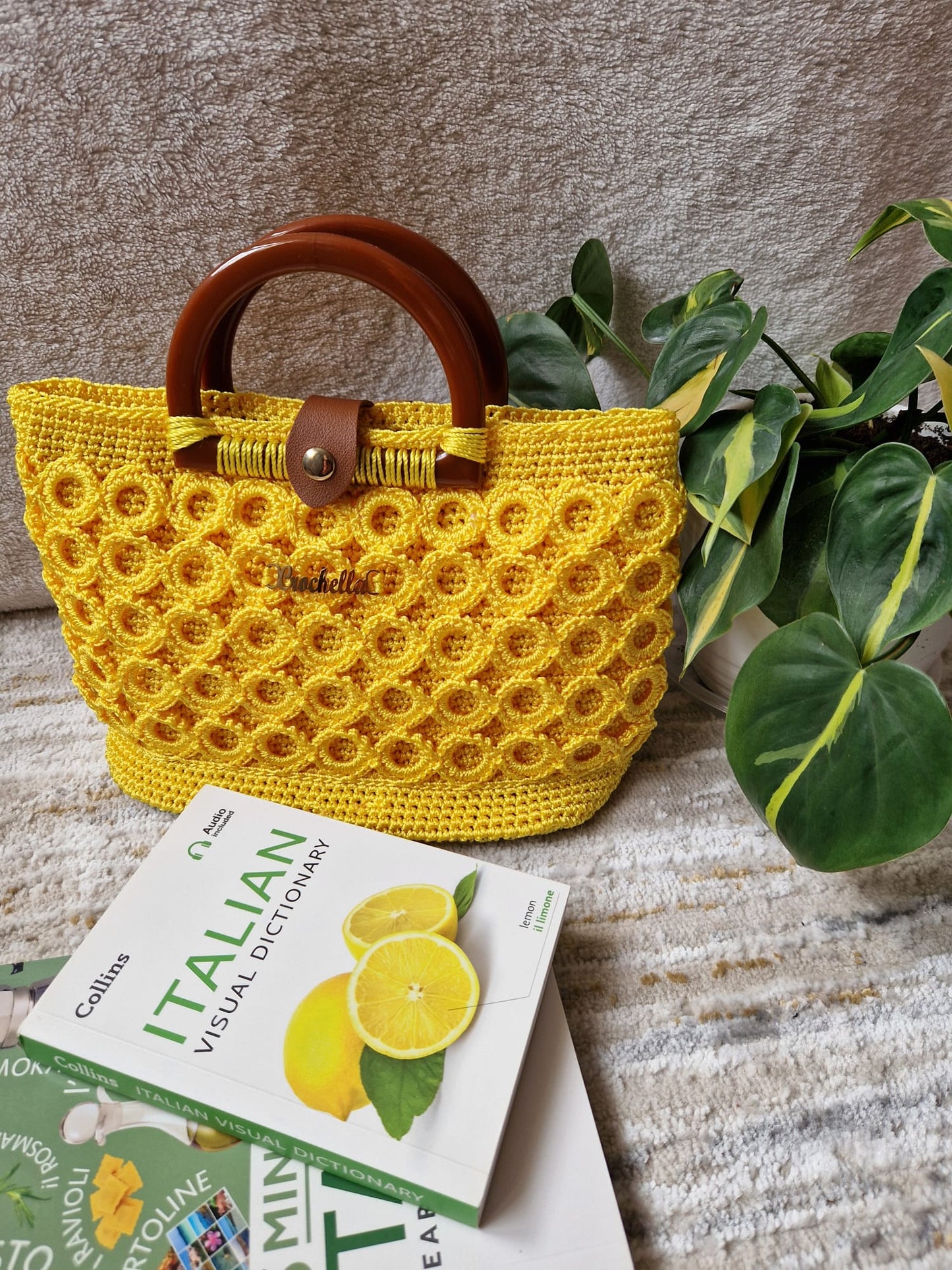 Sunburst Bloom Crochet Handbag in vibrant yellow, styled with an Italian dictionary and plant, highlighting its bold texture and round wooden handles