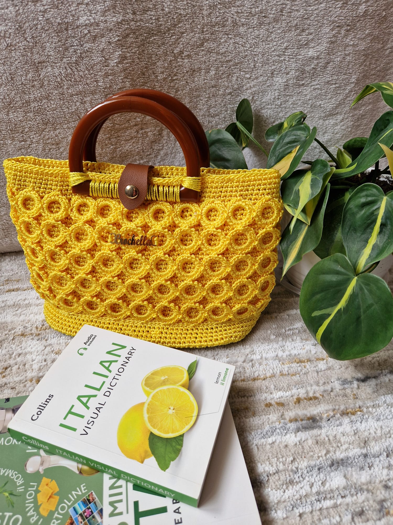 Sunburst Bloom Crochet Handbag in vibrant yellow, styled with an Italian dictionary and plant, highlighting its bold texture and round wooden handles