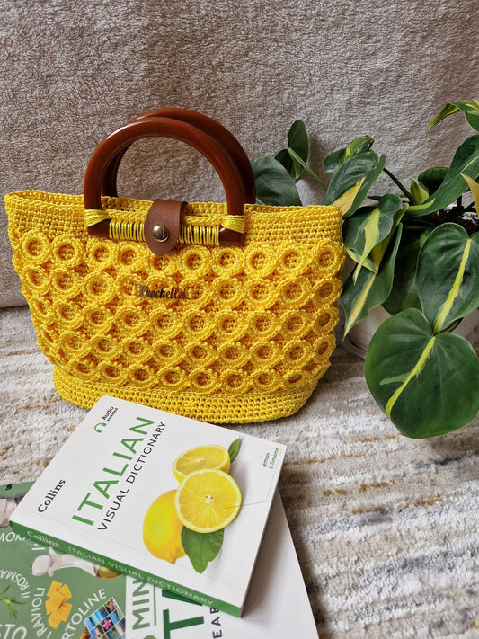 Sunburst Bloom Crochet Handbag in vibrant yellow, styled with an Italian dictionary and plant, highlighting its bold texture and round wooden handles