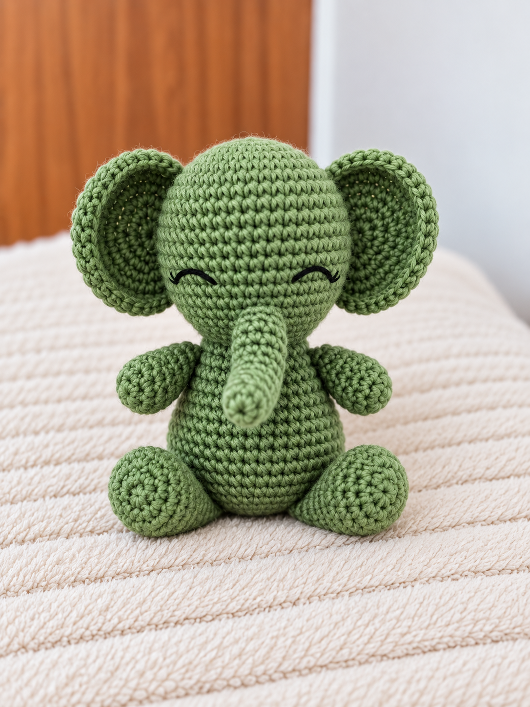 A green crocheted elephant toy sitting on a textured cream-colored blanket, with a wooden door and white wall in the background.