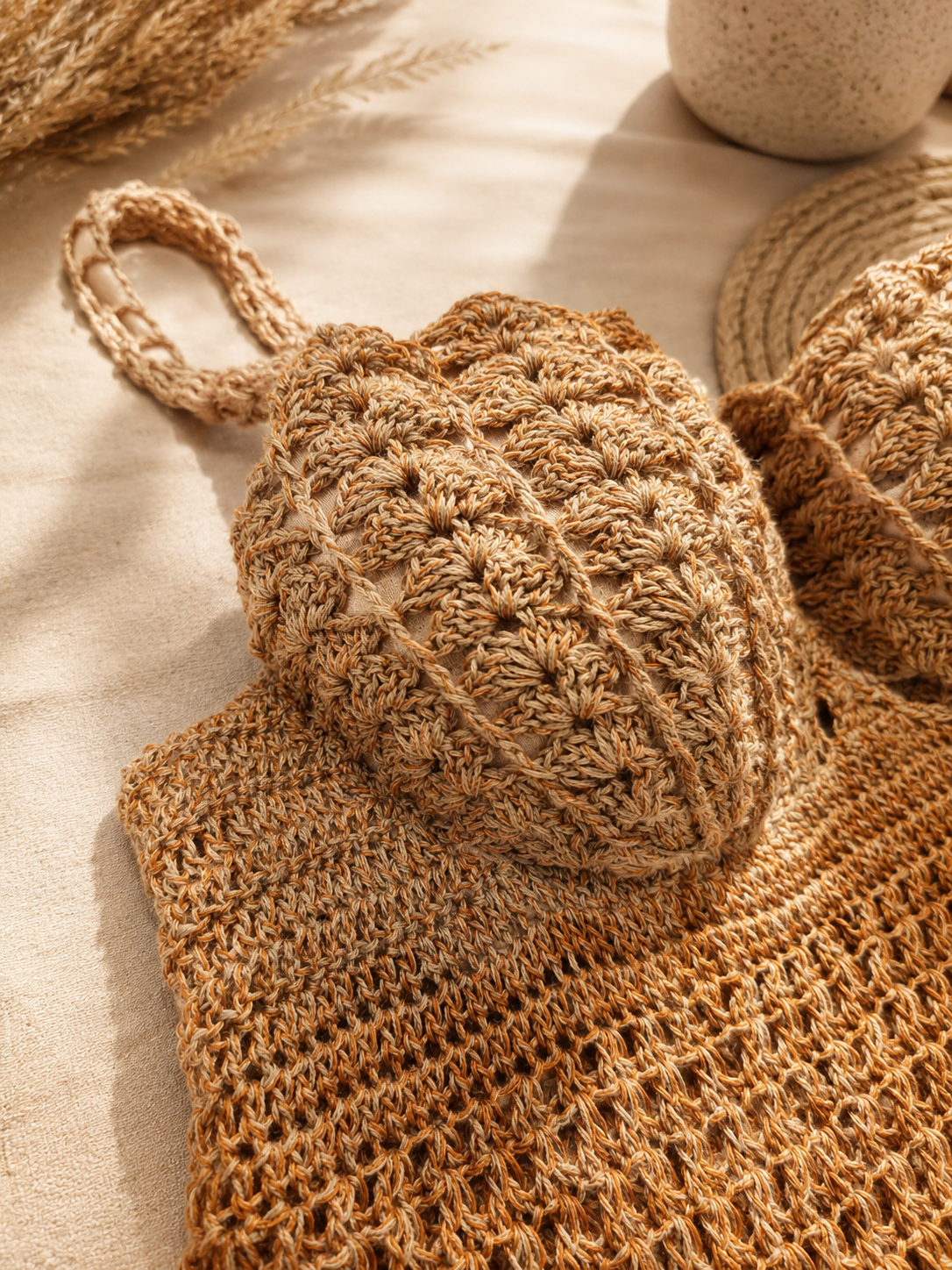 A close-up of a beige and brown crocheted knit bag with a textured pineapple pattern and a woven handle, styled on a neutral fabric surface with dried wheat and a woven mat in the background.