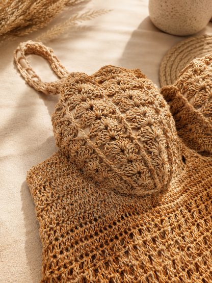 A close-up of a beige and brown crocheted knit bag with a textured pineapple pattern and a woven handle, styled on a neutral fabric surface with dried wheat and a woven mat in the background.