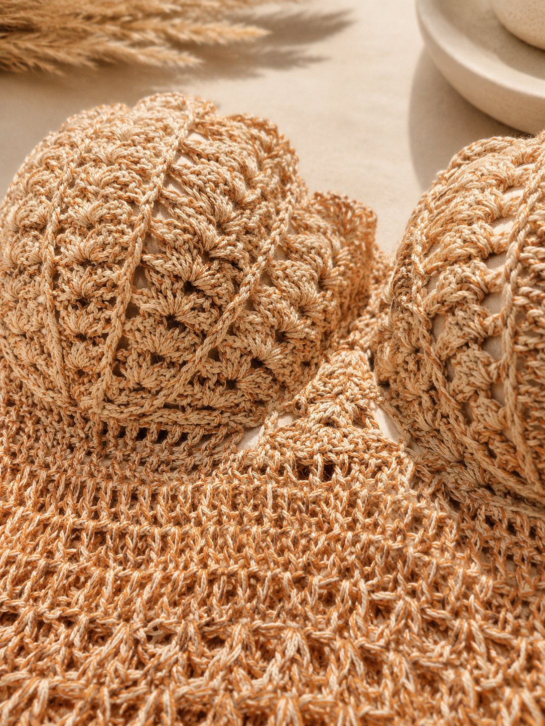 Close-up of a beige crochet knit fabric with textured floral and openwork patterns, shown on a light neutral surface with dried wheat and a ceramic dish in the background.