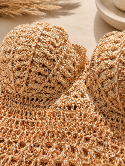 Close-up of a beige crochet knit fabric with textured floral and openwork patterns, shown on a light neutral surface with dried wheat and a ceramic dish in the background.