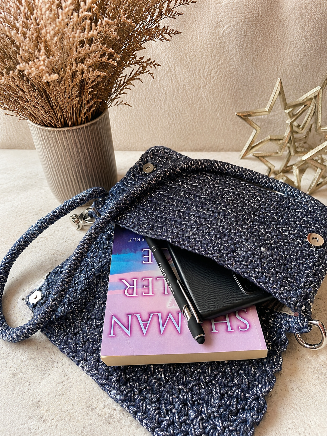 Blue knitted bag with a book and pen on a beige surface, with decorative elements in the background.