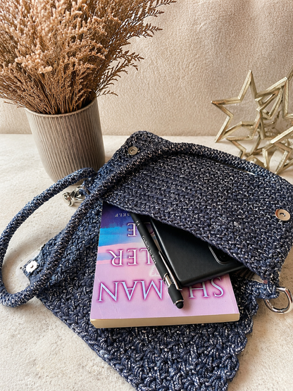 Blue knitted bag with a book and pen on a beige surface, with decorative elements in the background.