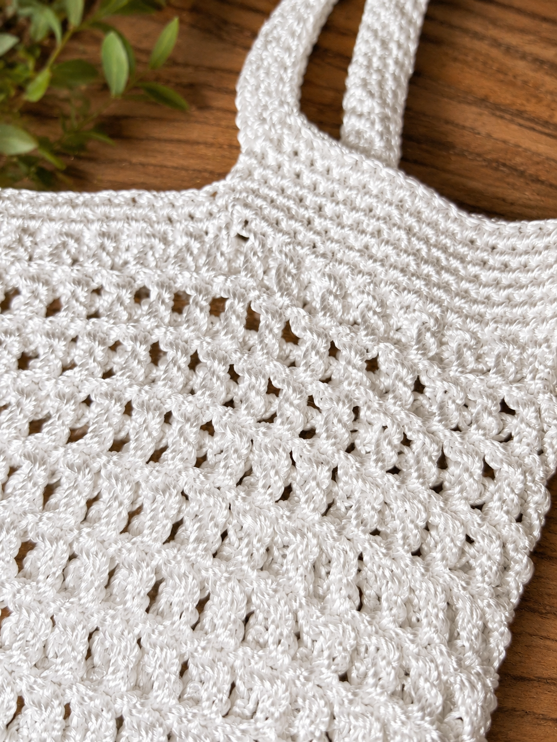Close-up of a white crochet bag on a wooden surface with a plant in the corner.