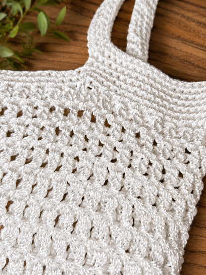 Close-up of a white crochet bag on a wooden surface with a plant in the corner.