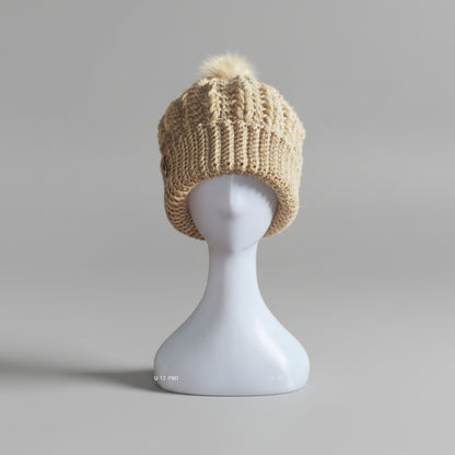Cozy beige crochet beanie with faux-fur pom-pom styled on a mannequin, set against a snowy winter backdrop.