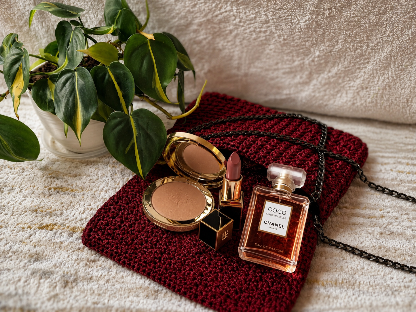 Chanel perfume bottle, makeup items, and a red textured pouch on a textured surface with a plant in the background.