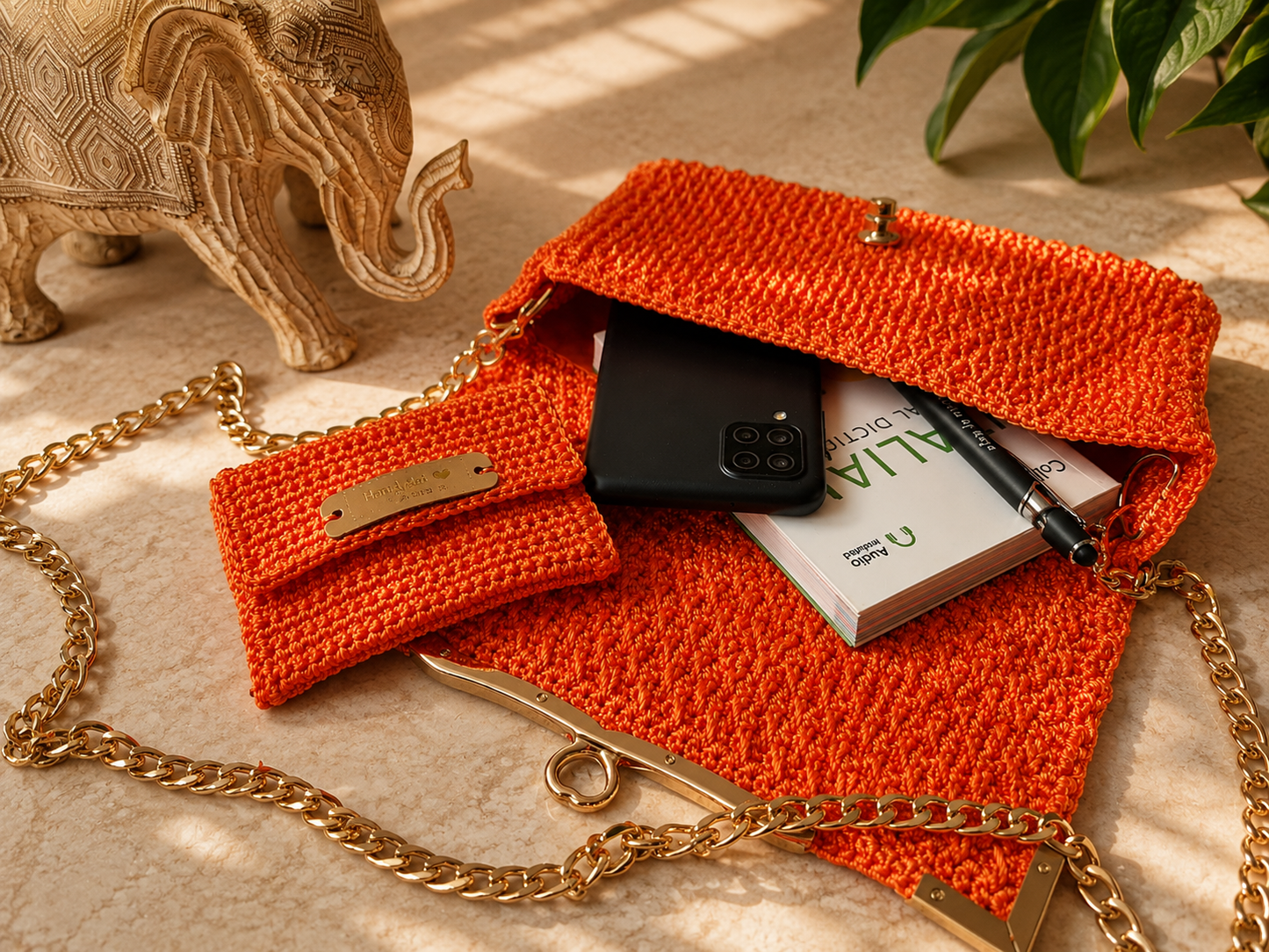 Orange crochet handbag with gold chain, phone, book, and pen on a beige surface with decorative elephant and plant.