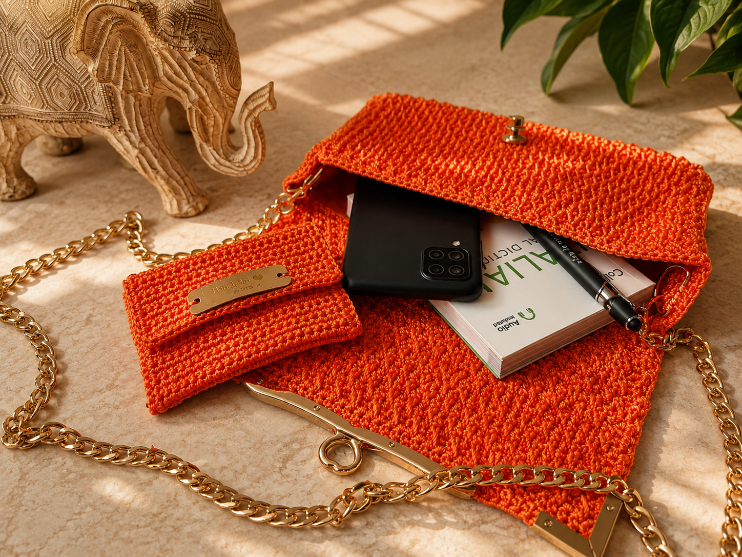 Orange crochet handbag with gold chain, phone, book, and pen on a beige surface with decorative elephant and plant.