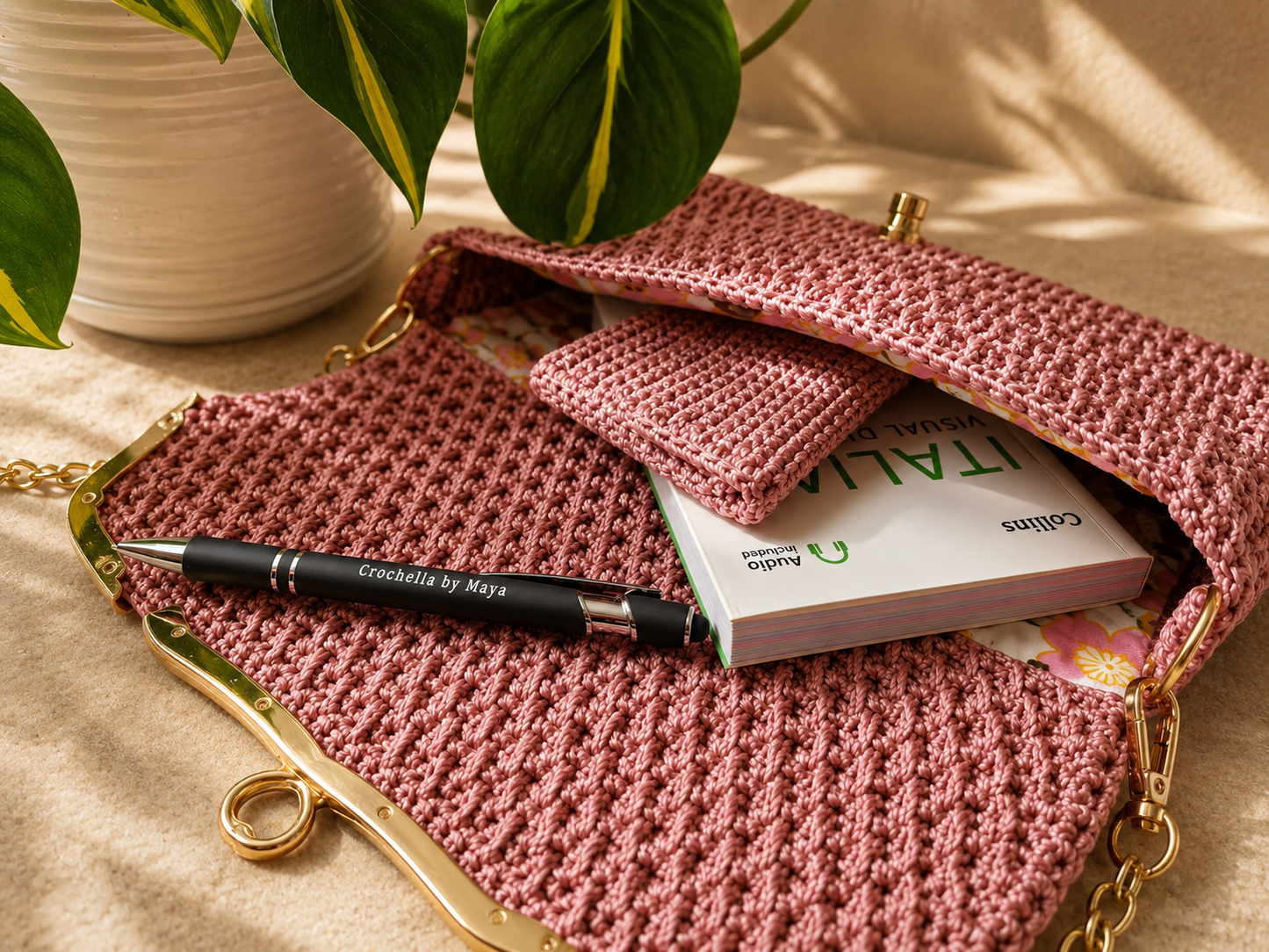 Pink crochet clutch with a pen and book on a beige surface with a plant in the background