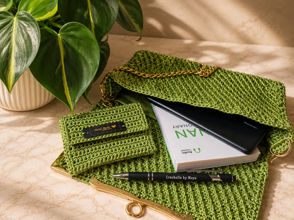 Green crocheted clutch with a book and pen on a beige surface, next to a plant.