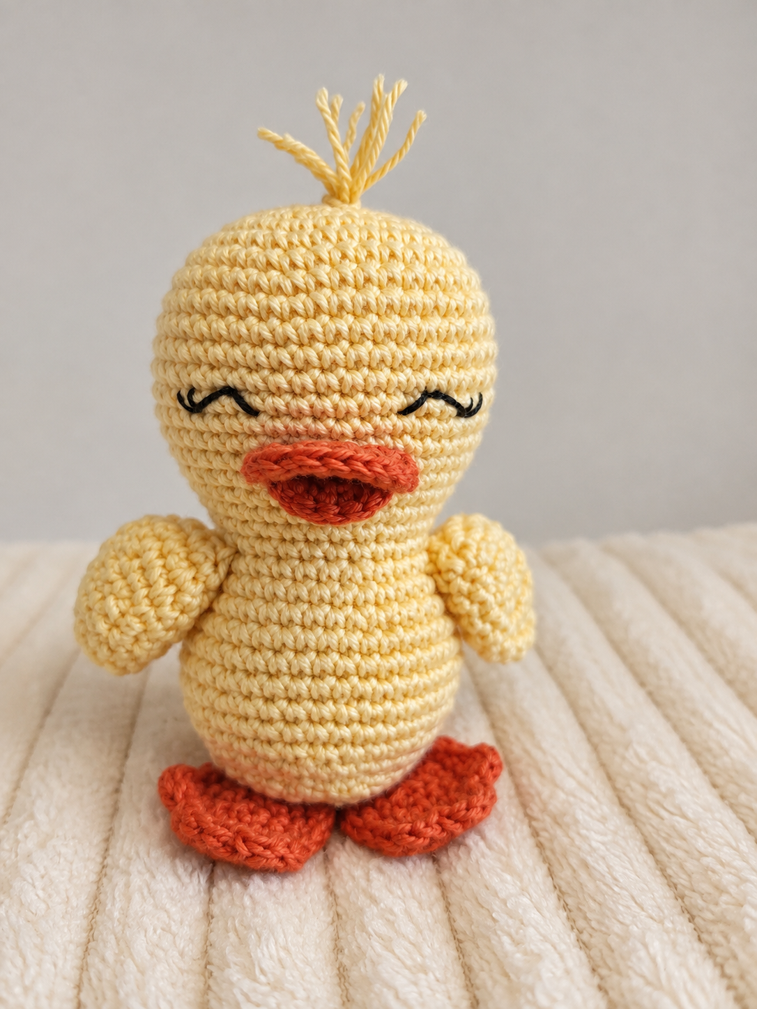 A crocheted amigurumi duck toy in pale yellow with orange-red beak and feet, photographed against a soft light gray background and cream ribbed fabric surface.
