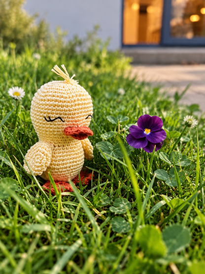 A crocheted yellow duck toy with an orange beak and feet sits in green grass beside a purple flower, with a blurred outdoor background.