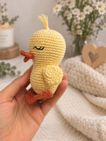 A hand holds a small crocheted yellow duck toy with orange beak and feet, set against a soft, cozy background with white daisies and a cream knit blanket.