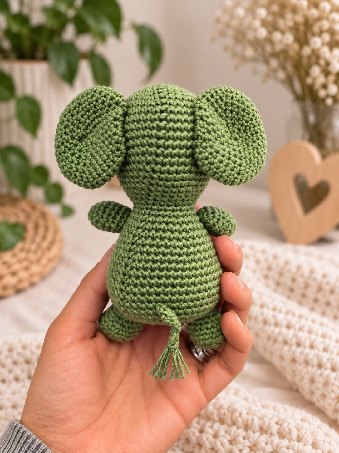 A small crocheted plush elephant toy in soft sage green is being held in a person's hand, with a cozy indoor background featuring a knit blanket, dried flowers, and a wooden heart decor piece.