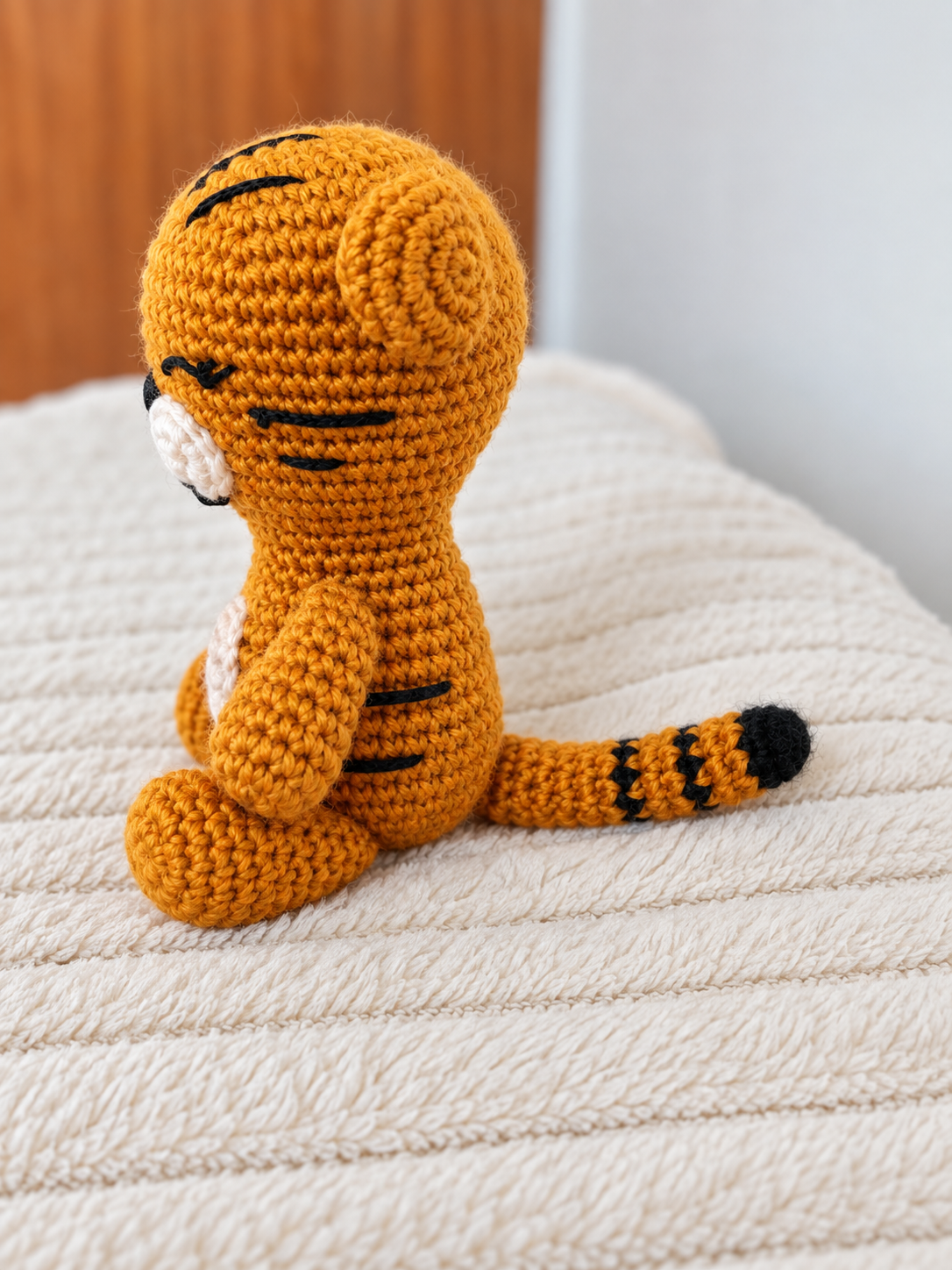 A crocheted tiger amigurumi toy sitting on a textured cream-colored blanket, with a wooden surface and white wall in the background.