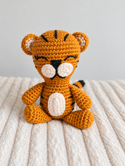 A crocheted tiger amigurumi toy sitting on a cream ribbed blanket against a light gray background.