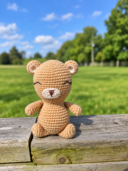 A small crocheted teddy bear toy sits on a weathered wooden surface outdoors, with a blurred green field and blue sky in the background.