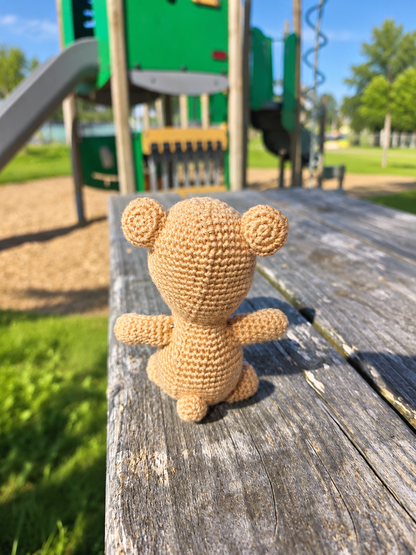 A small crocheted teddy bear toy sits on a weathered wooden picnic table in an outdoor playground setting, with a green play structure blurred in the background.