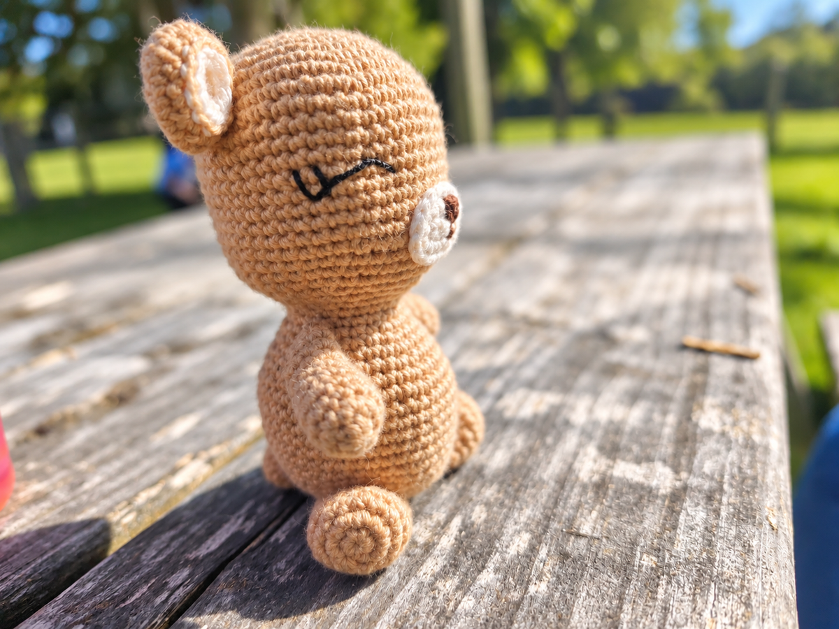 A small crocheted teddy bear toy sits on a weathered wooden picnic table outdoors, with a blurred green park background.